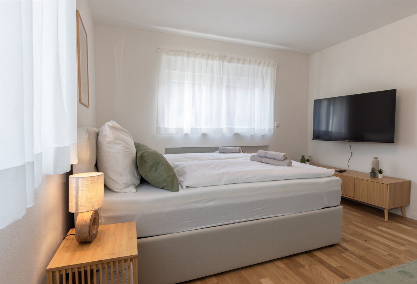Un dormitorio moderno con una cama grande, una mesita de noche con lámpara, un televisor montado en la pared y una ventana con cortinas translúcidas.