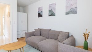 Una sala de estar moderna con un sofá gris, una mesa de centro de madera y un jarrón verde con plantas secas. Hay tres cuadros abstractos en la pared.