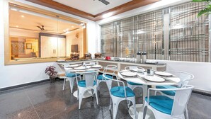 A modern dining area with white chairs and tables, a large window with blinds, and a view of the kitchen in the background. - Treebo Hr Grand (Tirupati)