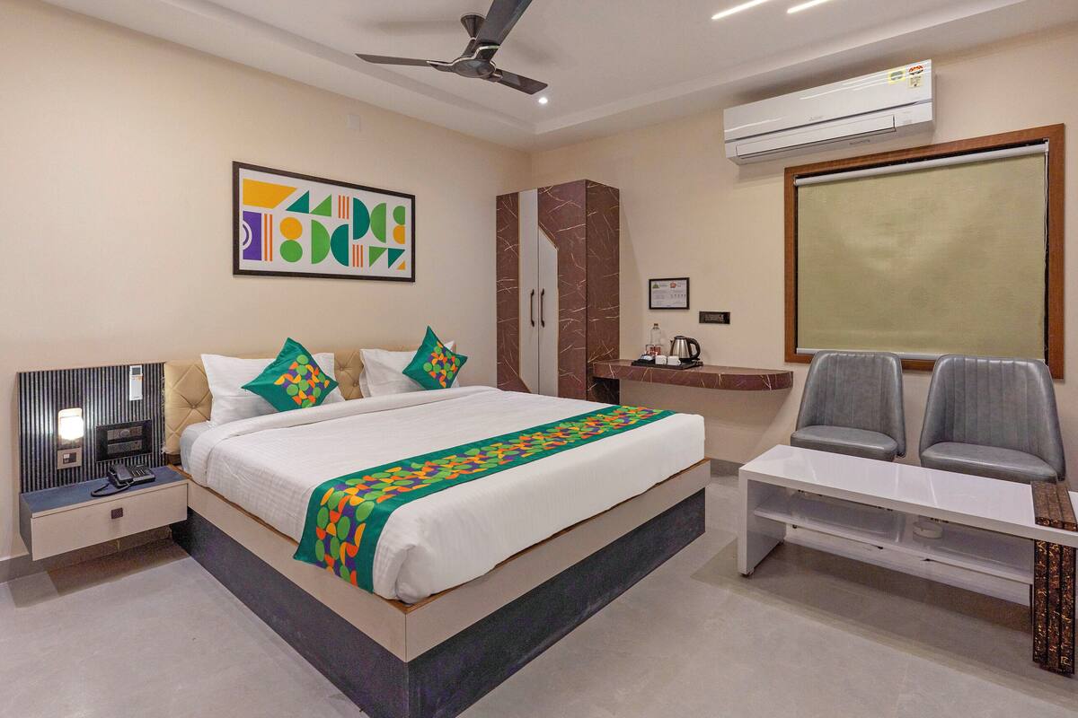 A modern hotel room with a bed, a desk, two chairs, a ceiling fan, an air conditioner, and a framed abstract artwork on the wall.