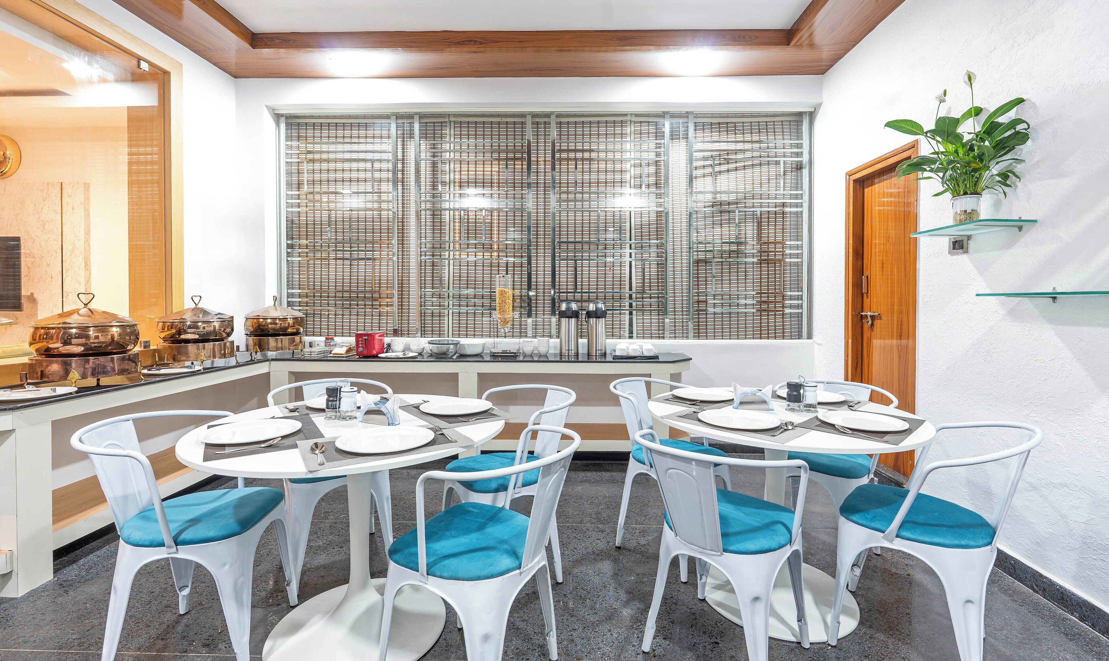 Un comedor con mesas redondas y sillas blancas, un bufé con diversos alimentos, y un ventanal con persianas.