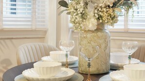 Une table à manger dressée avec de l’assiette blanche, des bols et des verres à vin, agrémentée d’un vase de fleurs.