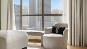A modern hotel room with a large window, a grey armchair with a black cushion, and a white bed. - 2BR Marina View Steps from the Beach (Dubai)