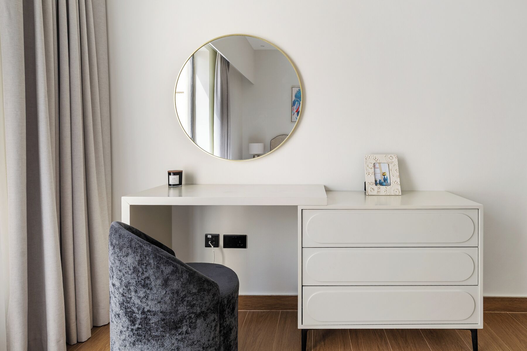A modern bedroom with a white dresser, a round mirror, a small bedside table with a candle, and a chair.