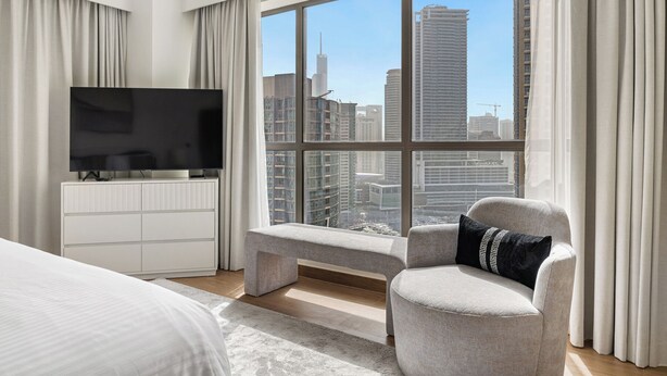 A modern bedroom with a large window, a flat-screen TV, a bed, a bench, and a chair. - 2BR Marina View Steps from the Beach (Dubai)