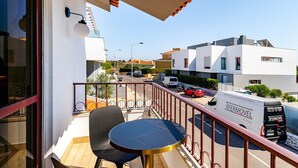 Een balkon met een ronde tafel en twee stoelen, met uitzicht op een straat met geparkeerde auto's en gebouwen.