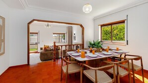 Een eethoek met een houten tafel gedekt voor vier personen, omringd door rieten stoelen. Een groot raam biedt uitzicht op groen buiten.