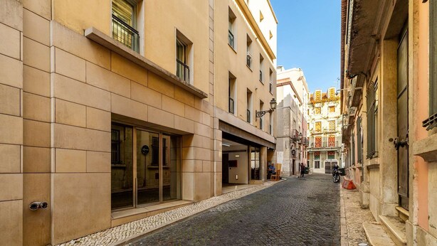 A narrow cobblestone street flanked by buildings with large windows and doors, suggesting a historic urban setting. - Modern Flat in Central Lisbon (Lisbon)