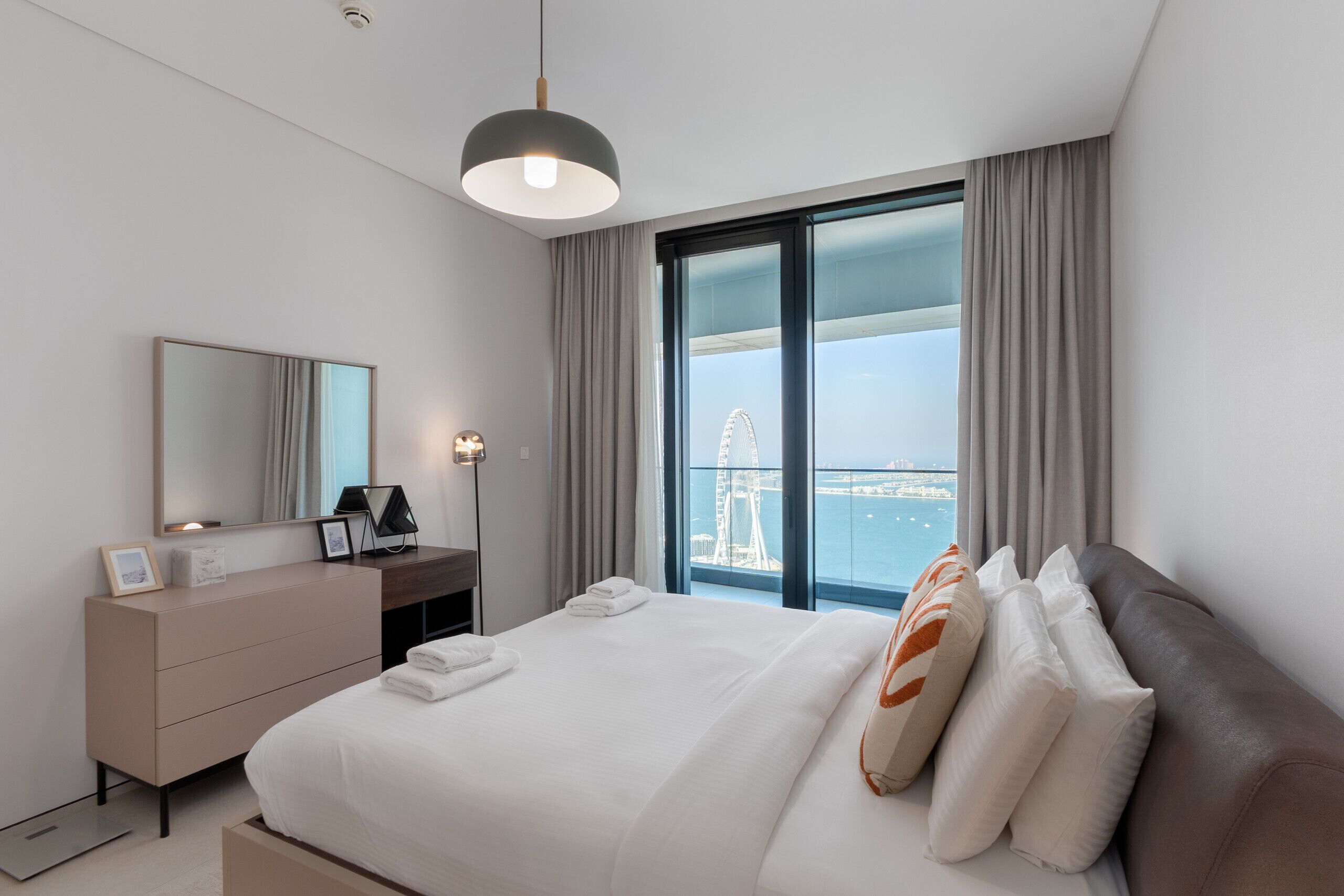 A modern hotel room with a large bed, a dresser, a desk, a mirror, and a view of a Ferris wheel.