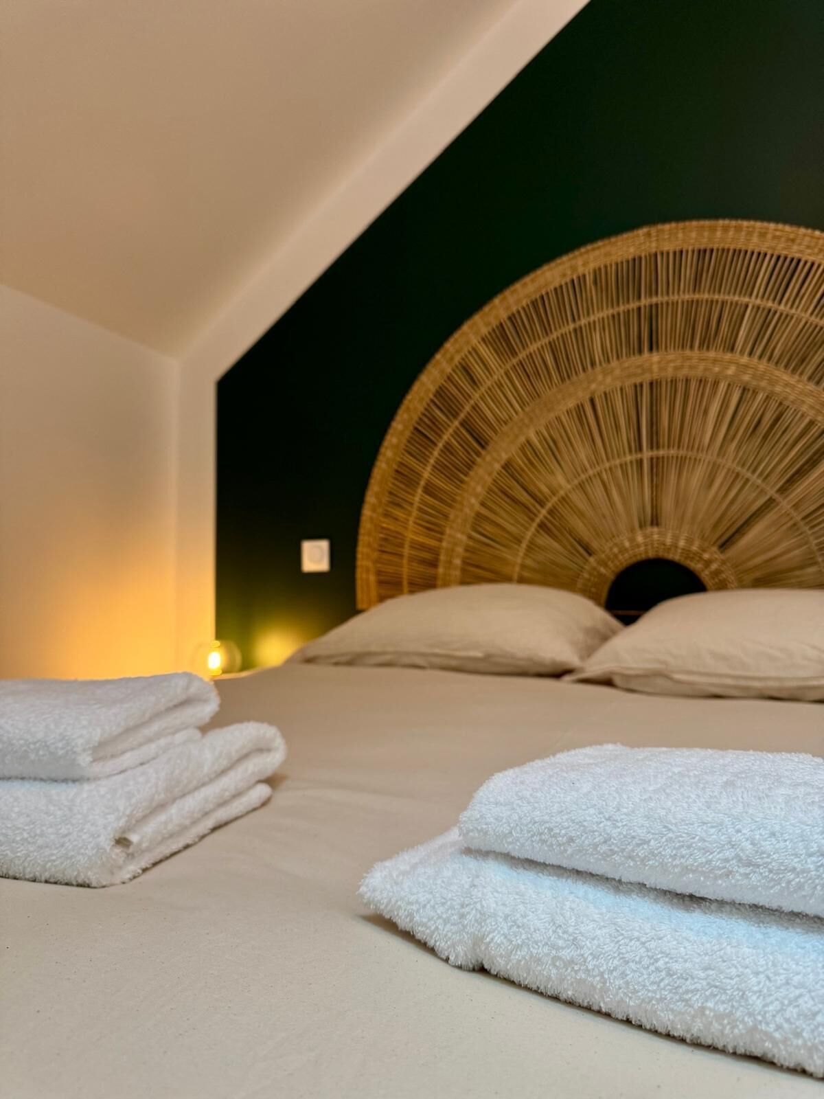 A room with a bed featuring a circular woven headboard, neatly folded white towels on the bed, and a small lit candle on the nightstand.