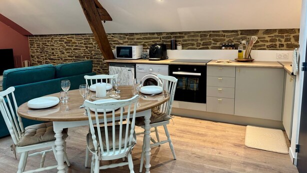 Un coin repas chaleureux avec une table en bois dressée pour deux personnes, des chaises blanches et un mur en pierre en arrière-plan.