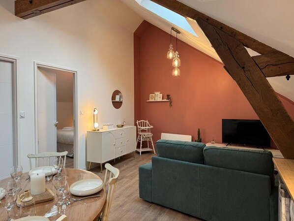 Una sala de estar acogedora con un sofá verde, una mesa de comedor de madera para cuatro personas, un armario blanco y un televisor.