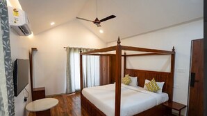 Uma cama de dossel com estrutura de madeira e roupa de cama branca, um ventilador de teto, uma TV fixada na parede e uma mesa redonda.
