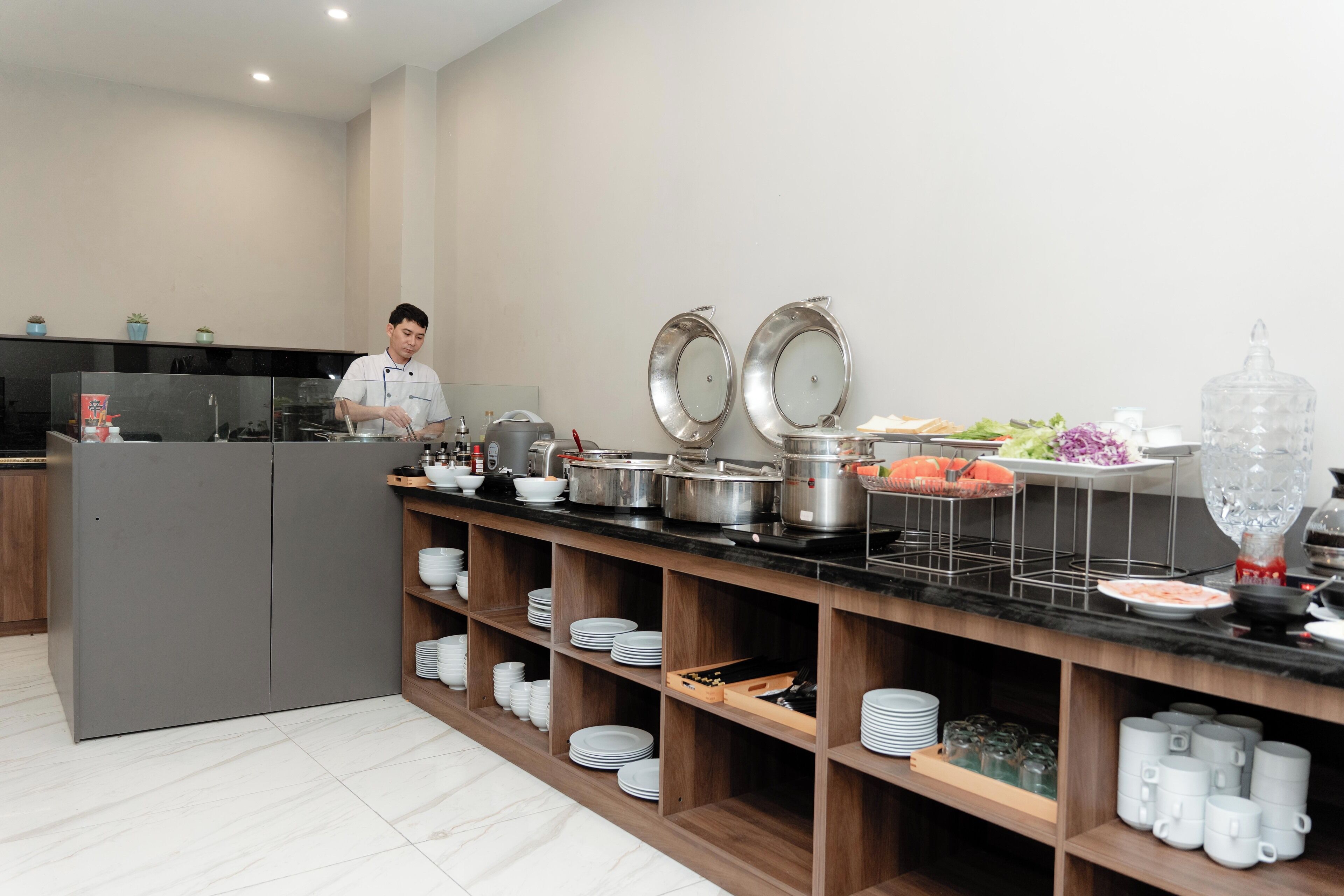 Una cocina moderna con un chef preparando comida, equipada con electrodomésticos de acero inoxidable, repisas de madera con platos y una variedad de alimentos.