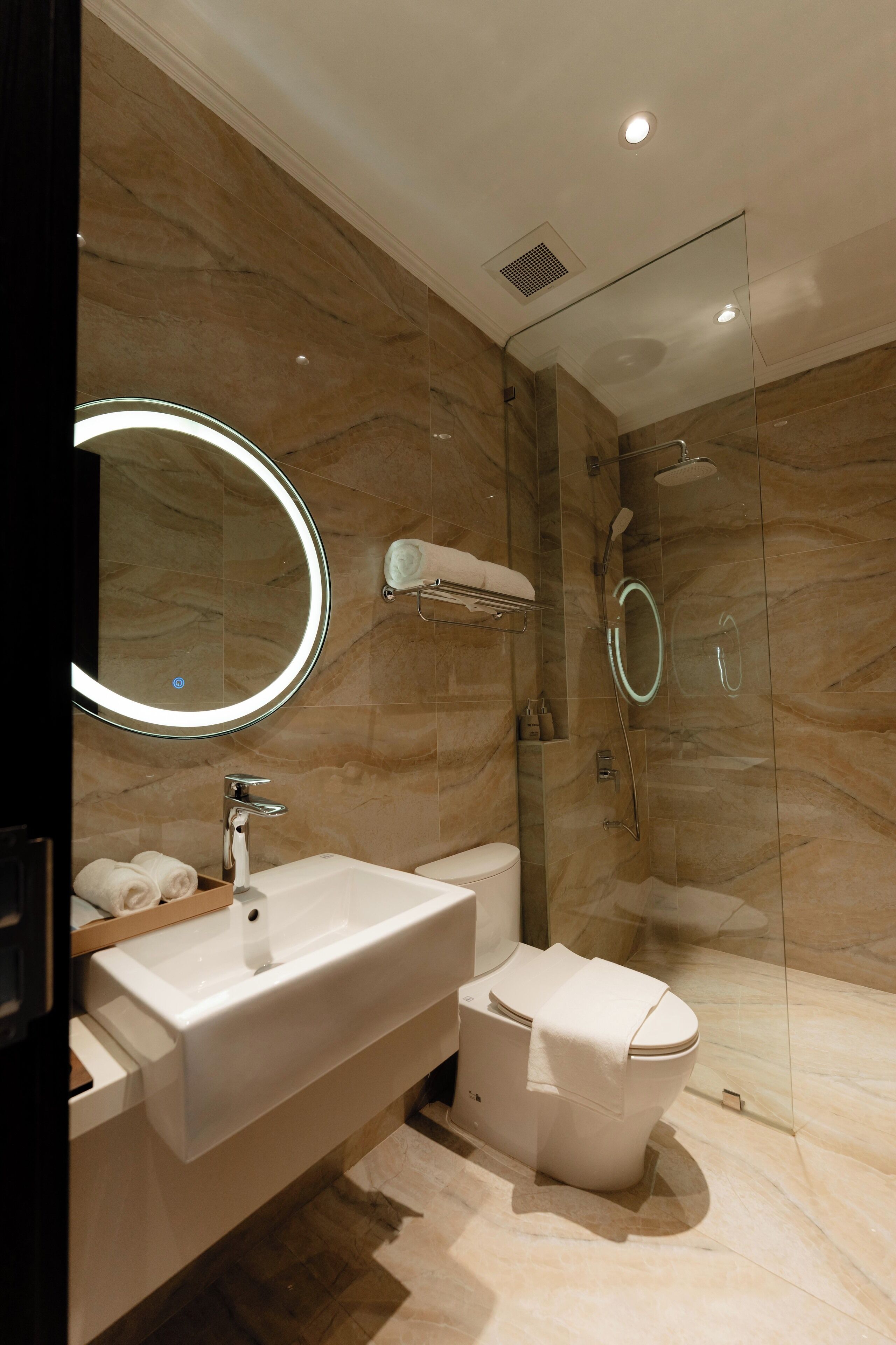 Un baño moderno con pared de mármol, un espejo redondo, un lavamanos blanco, un inodoro y una zona de ducha con mampara de vidrio.