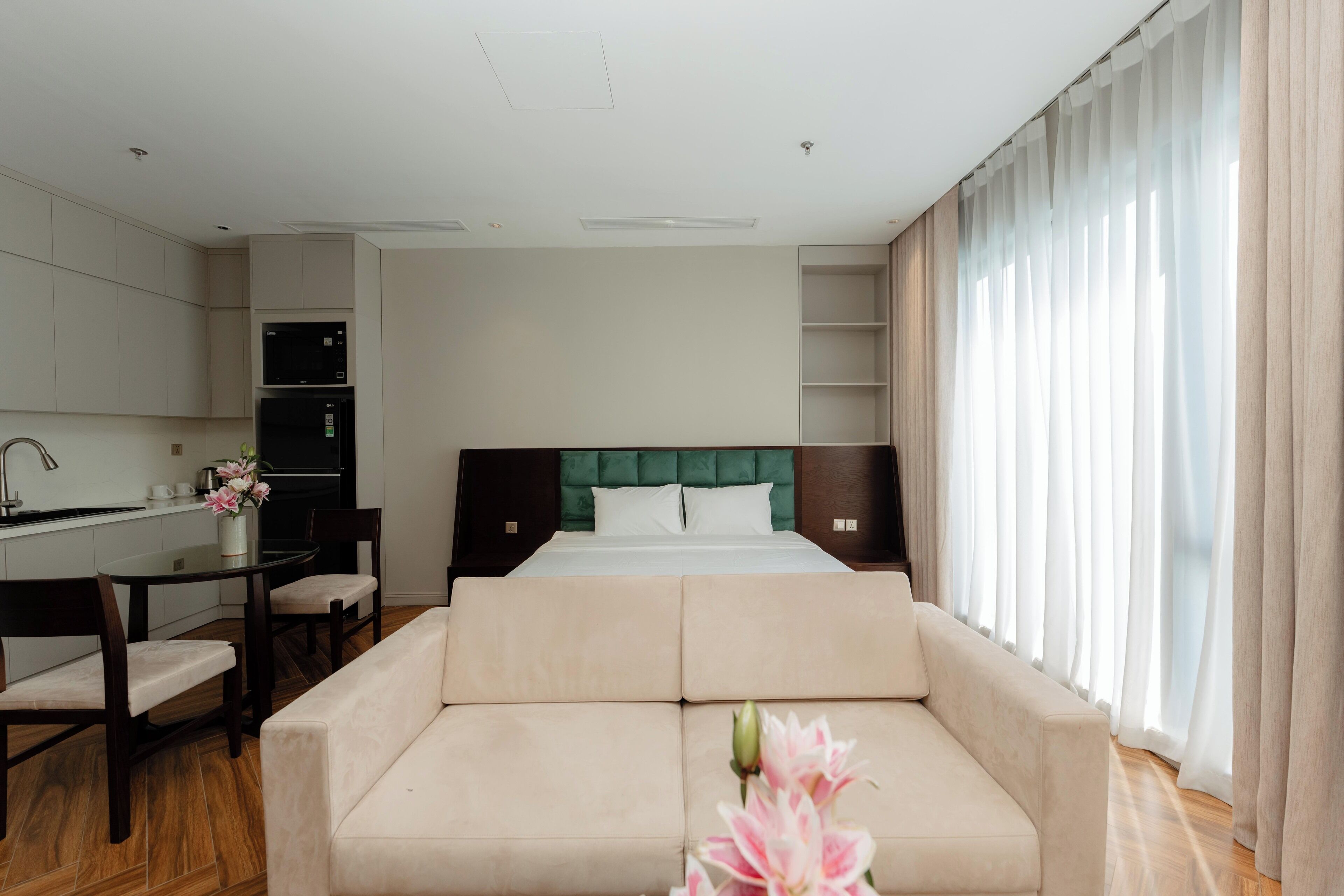 A hotel room with a bed, a sofa, a dining table, chairs, a kitchen area, and a large window with curtains.