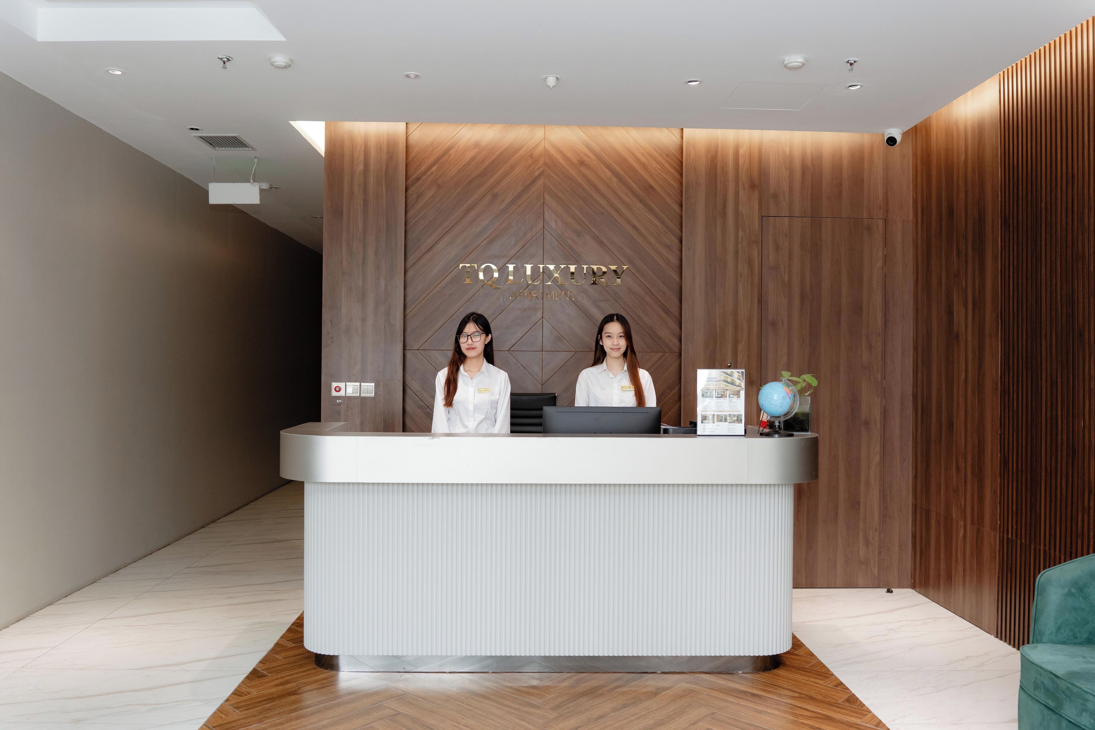 Een receptiebalie met een houten paneel als achtergrond, waarop de naam 'TO LUXURY' duidelijk staat vermeld. Achter de balie zitten twee receptionisten, met een computer en een kleine wereldbol op de balie zelf.