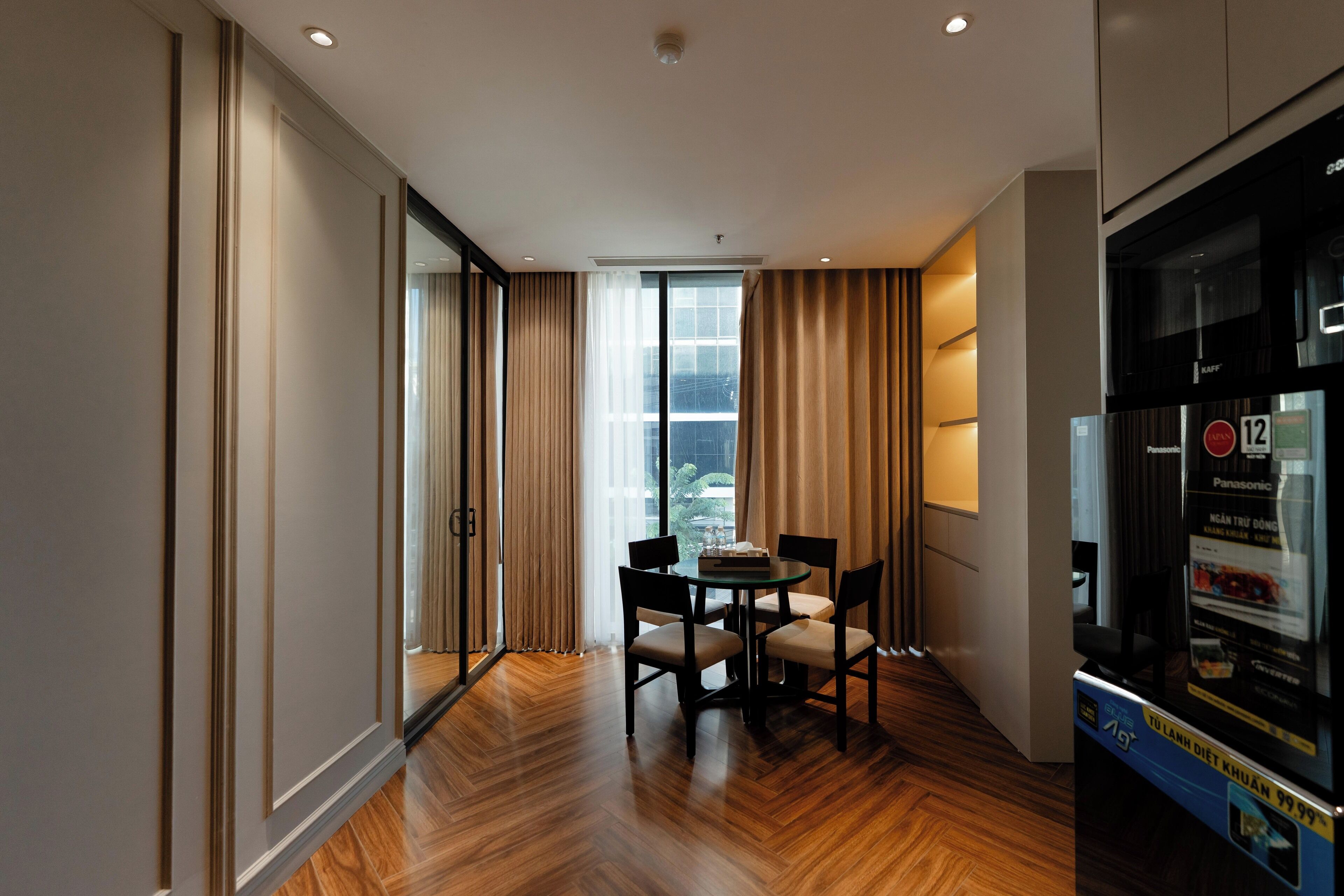 Ein modernes Hotelzimmer mit Holzboden, einem Esstisch mit Stühlen, einem großen Fenster mit Vorhängen und einer Minibar.