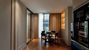 Ein modernes Hotelzimmer mit Holzboden, einem Esstisch mit Stühlen, einem großen Fenster mit Vorhängen und einer Minibar.