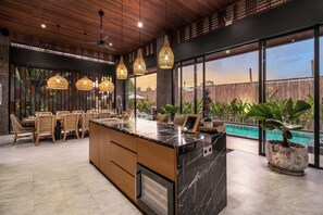 A modern kitchen with a marble island, wicker furniture, and a view of a pool. - 12 Bedroom Myosotis VVV (Ubud)