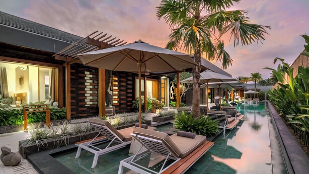 A modern outdoor pool area with lounge chairs and umbrellas, surrounded by lush greenery and a wooden fence. - 12 Bedroom Myosotis VVV (Ubud)