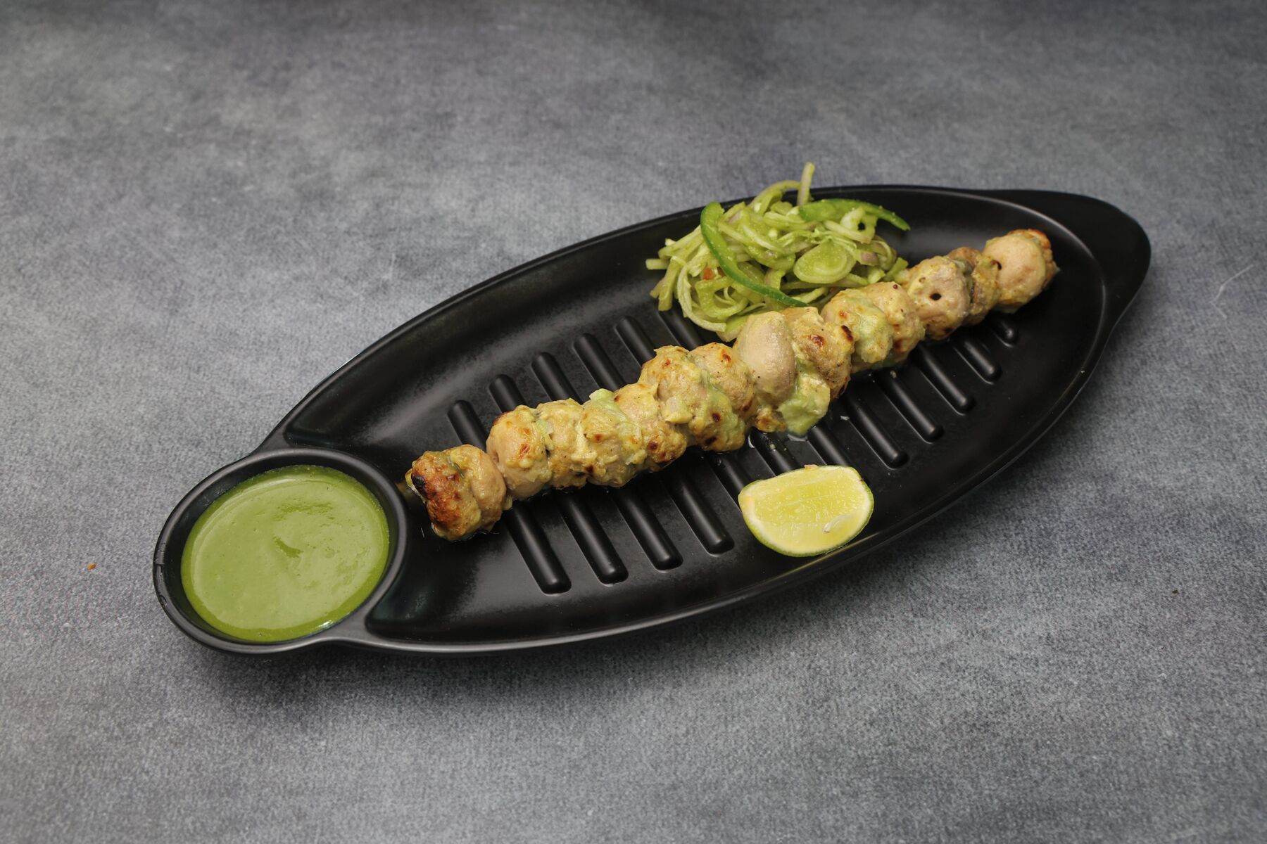 A grilled meat skewer with vegetables and a lime wedge on a black plate.