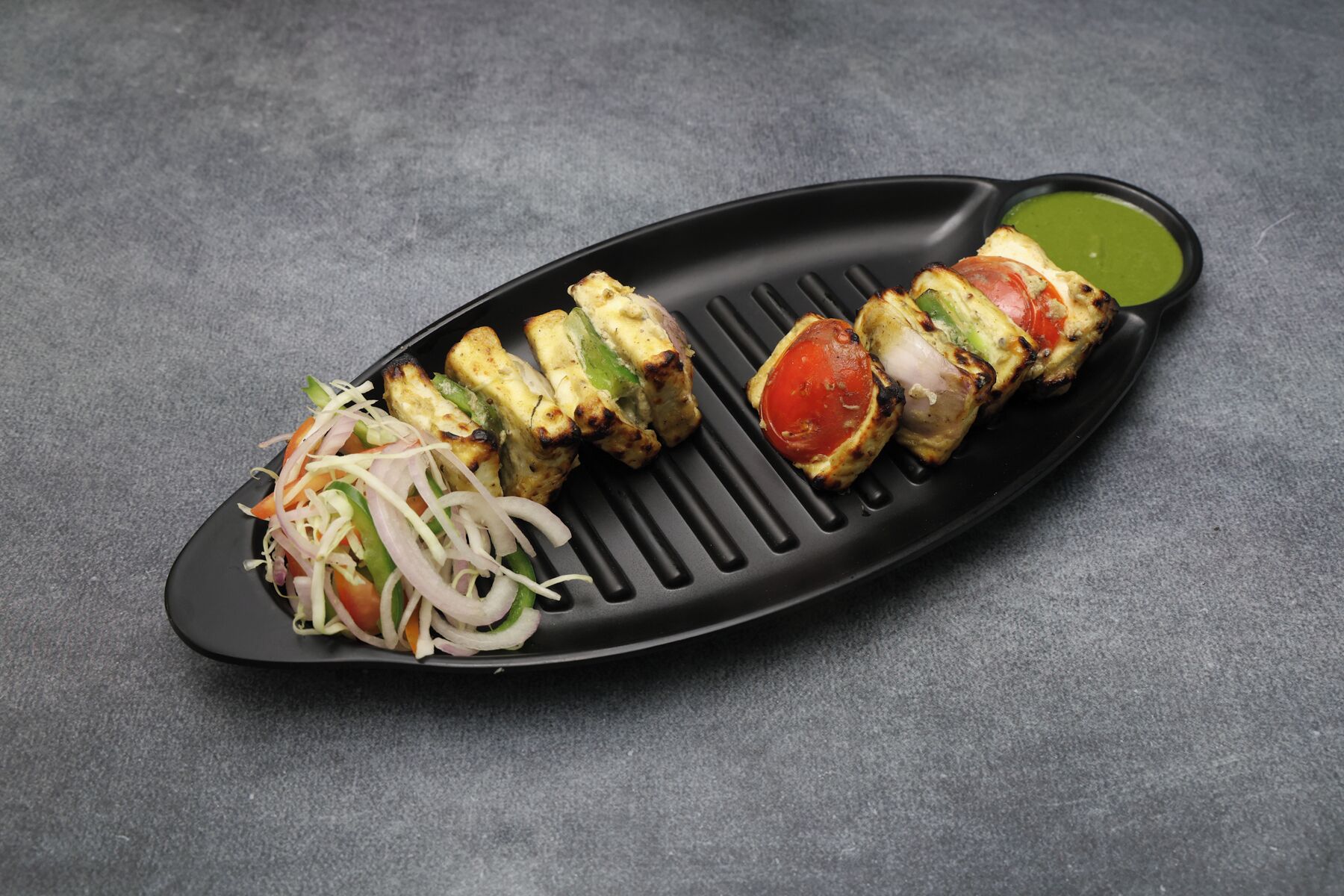 A black plate with grilled chicken skewers, a side salad, and a green sauce.