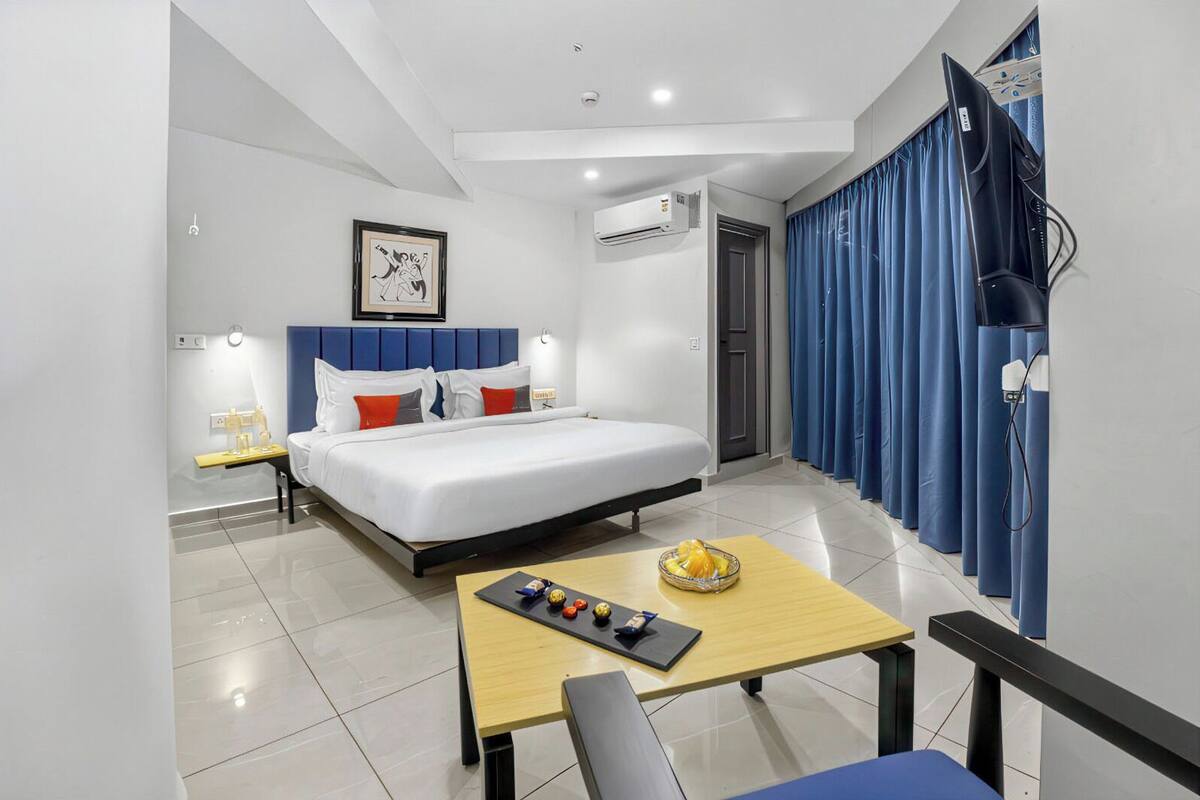 A hotel room with a bed, bedside table, lamp, air conditioning unit, curtains, and a TV.
