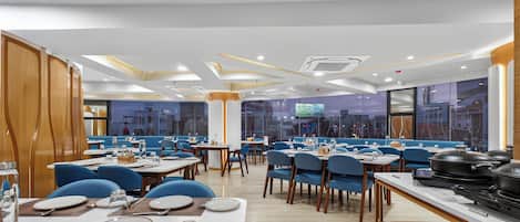 A modern restaurant with a clean, organized layout featuring blue chairs, white tables, and a central kitchen area.