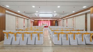 Une salle de conférence avec des rangées de chaises, une scène surmontée d’un fauteuil doré et un lustre.