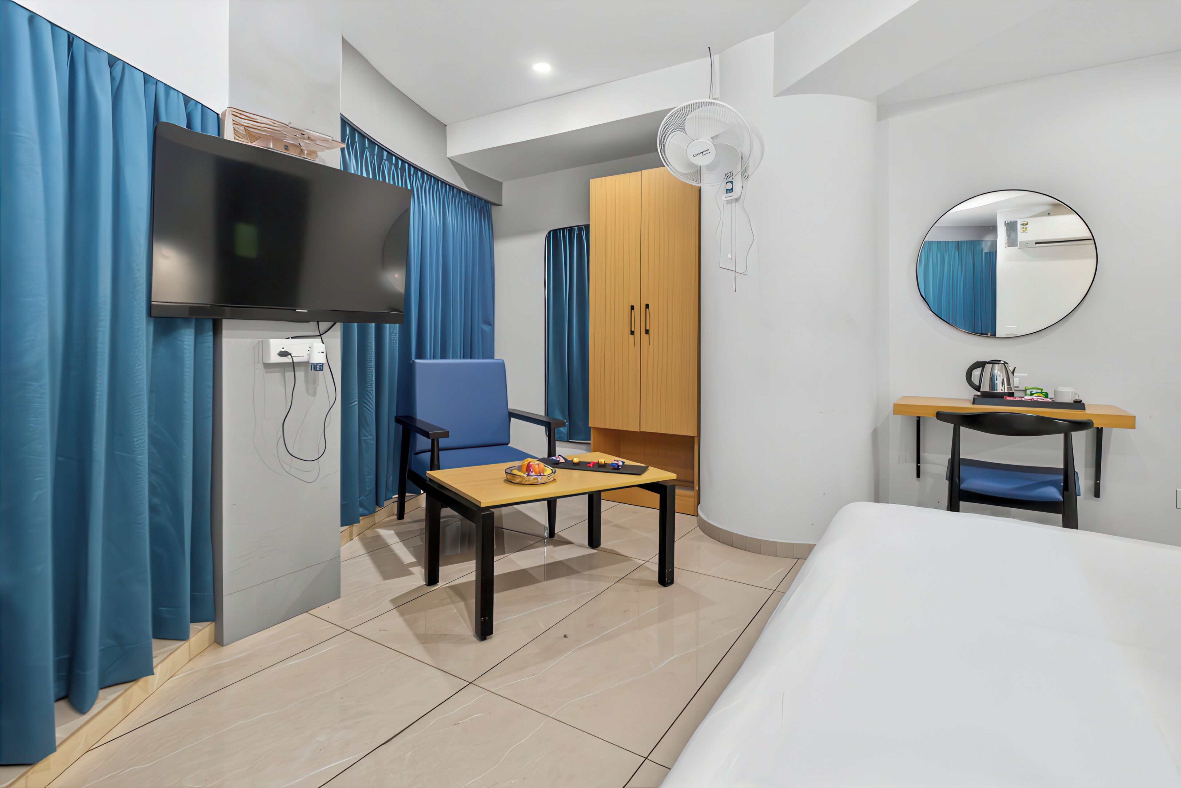 A hotel room with a bed, a desk with a chair, a television, a wardrobe, a mirror, and a fan.