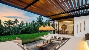 Un espacio moderno al aire libre con una pérgola de madera, una zona de comedor con paredes de vidrio y vistas a un jardín exuberante.