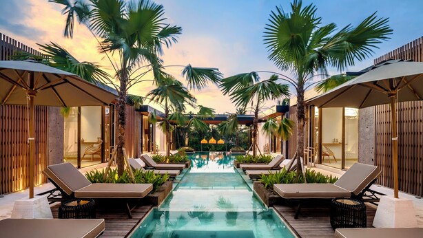 Una piscina con sillas de descanso y palmeras, rodeada de cercas de madera y arquitectura moderna.