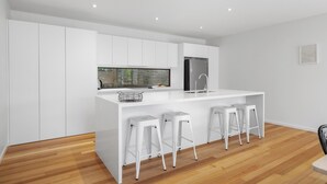 Une cuisine moderne avec des meubles blancs, un îlot central doté de tabourets, un réfrigérateur en acier inoxydable et une fenêtre donnant sur un espace verdoyant.