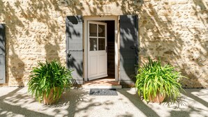 Un bâtiment en pierre avec une porte blanche, deux plantes en pot et un paillasson en métal.