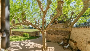 Une cour bordée d’arbres, avec un mur en pierre, une zone pavée et deux fauteuils de jardin.
