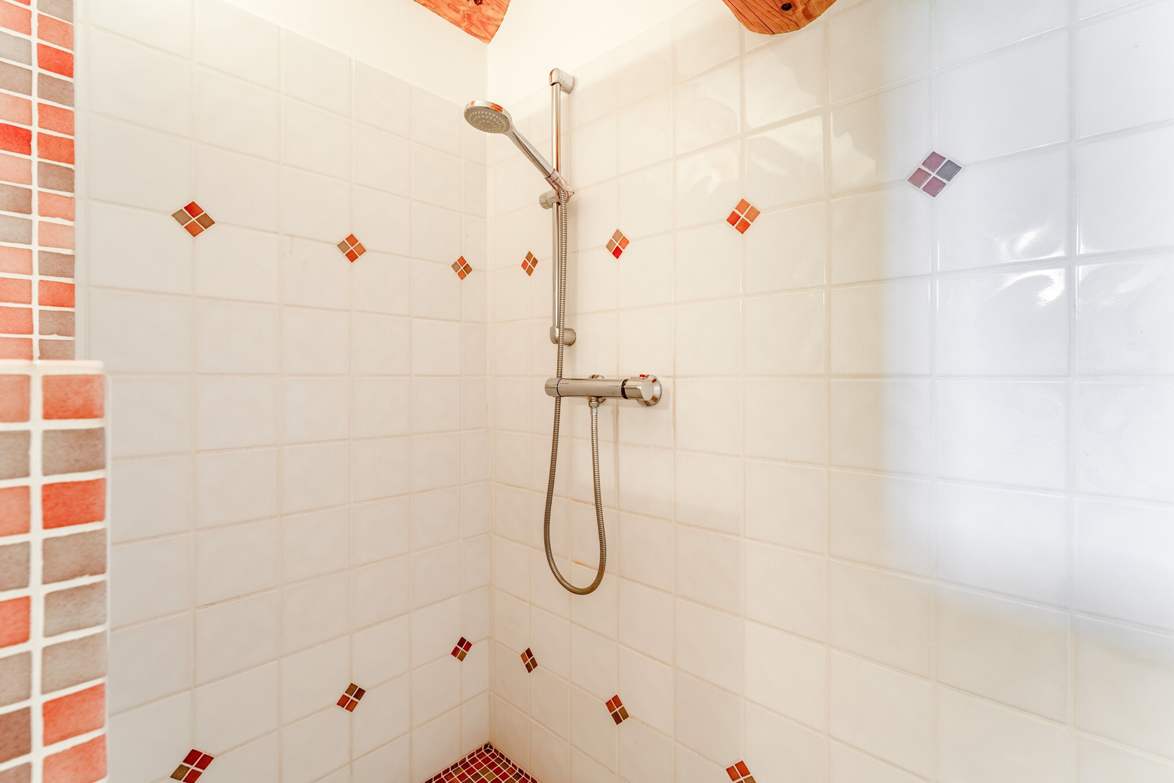 A shower with a handheld showerhead and a wall-mounted faucet, tiled walls with a pattern of small squares, and a corner with a red and white checkered floor.