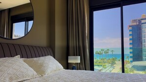 A hotel room with a large bed, a bedside table with a lamp, a view of the beach, and a tall building in the background. - Apartment 1+1 Qoople Excellence B19 100 m from the sea (Alanya)