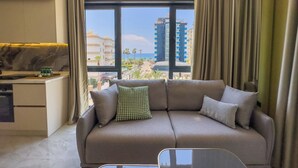 A modern hotel room with a sofa, a small table, and a view of the cityscape through large windows. - Apartment 1+1 Qoople Excellence B19 100 m from the sea (Alanya)