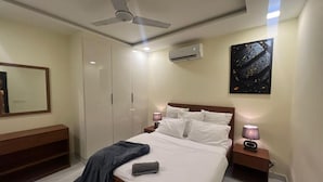 Une chambre moderne comprenant un lit, des tables de chevet, un ventilateur de plafond, un appareil de climatisation, un banc et une œuvre d’art accrochée au mur.