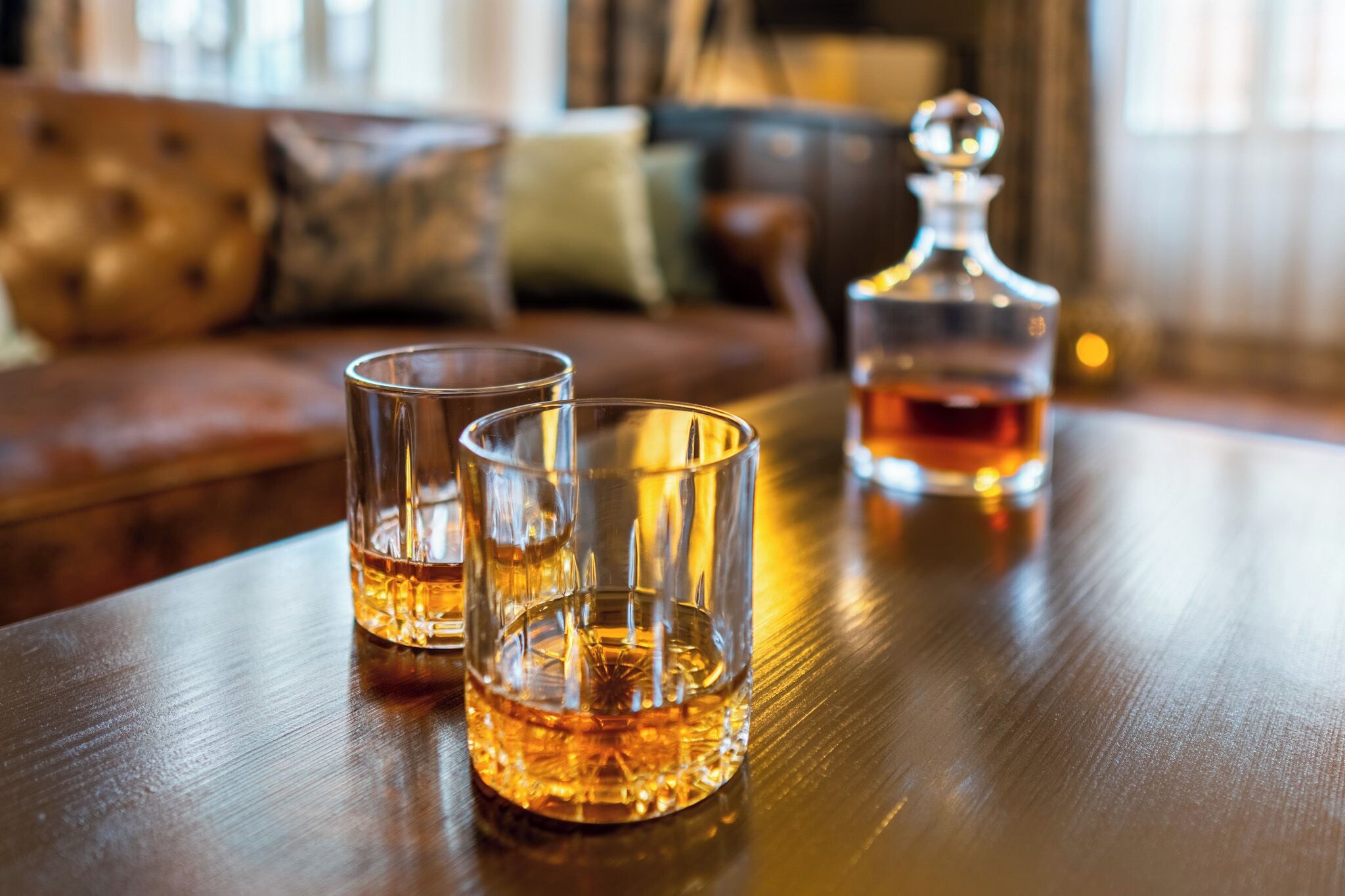 Three glasses of whiskey on a wooden table with a decanter in the background.