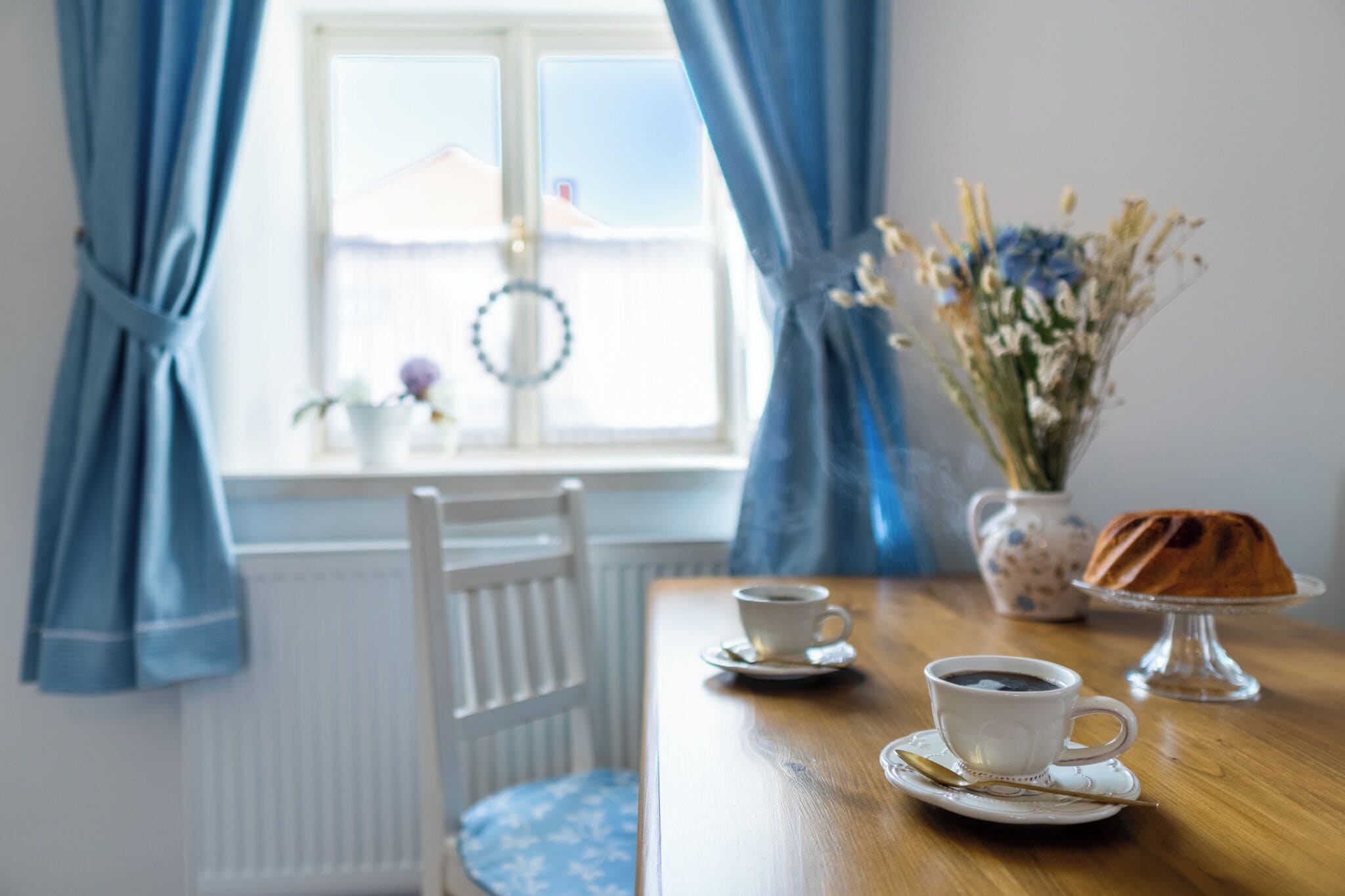 A dining table with two cups of coffee, a cake on a stand, and a vase of flowers.