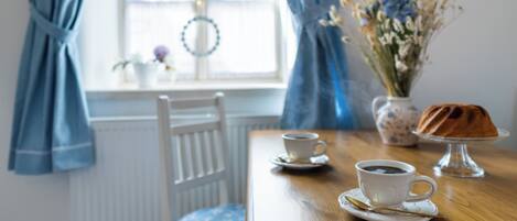 A dining table with two cups of coffee, a cake on a stand, and a vase of flowers.