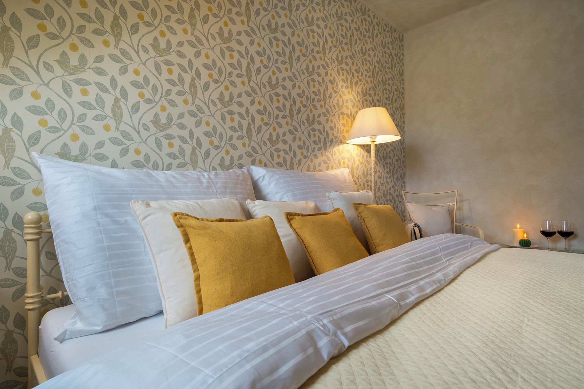 Una cama bien hecha con almohadas blancas y mostaza, una lámpara de noche y dos copas de vino sobre una mesita de noche.