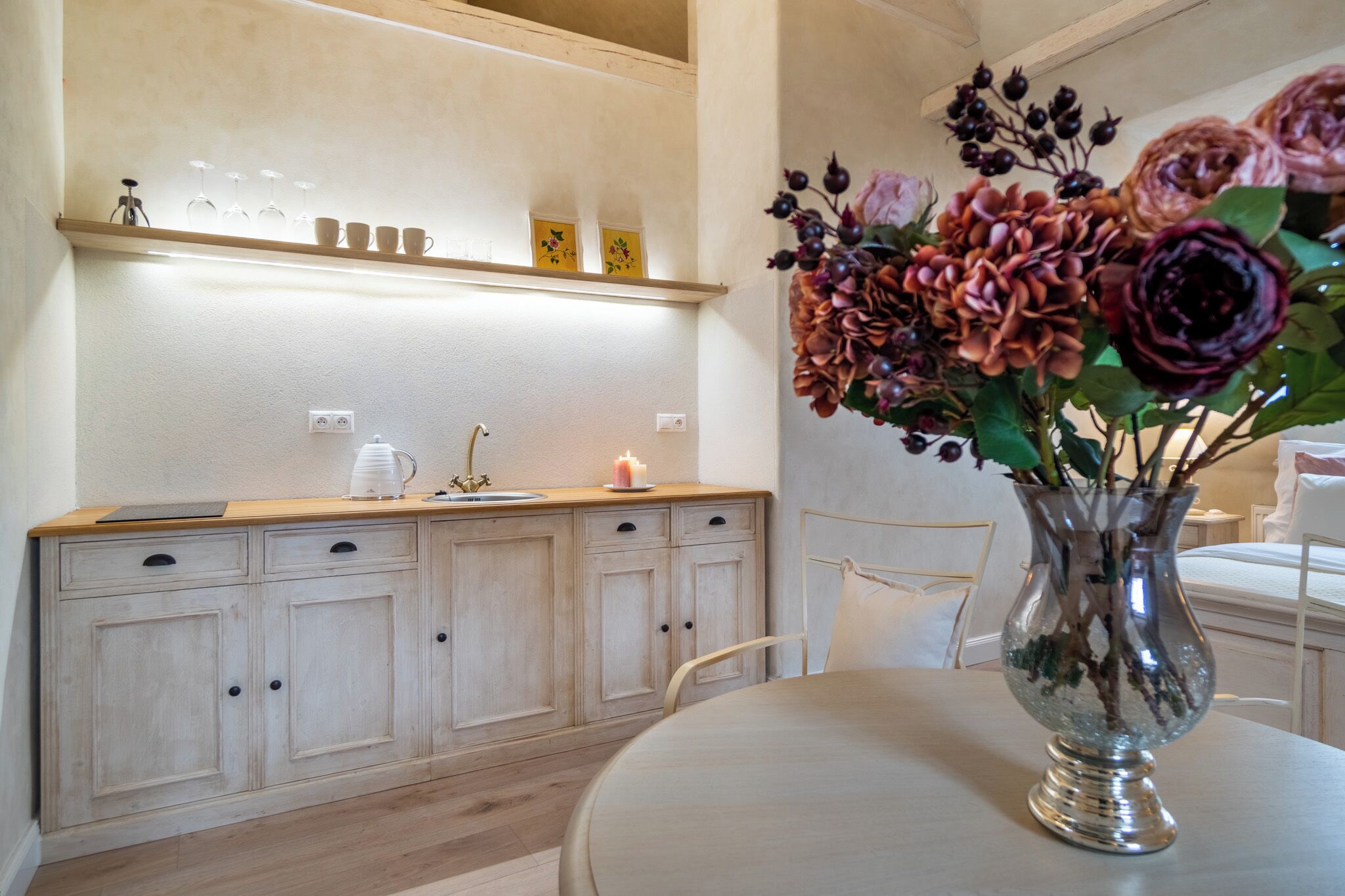 A dining area with a round table, a vase with flowers, a shelf with glasses and a candle, and a wooden cabinet with drawers.