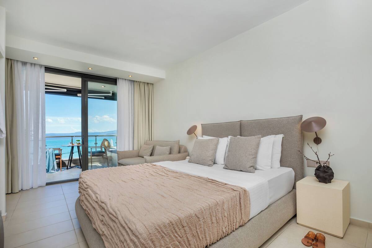 A bedroom with a large bed, a sofa, a small table, and a view of the sea.