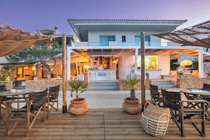 Terraza de madera con mobiliario exterior, plantas en macetas y un edificio con zona de bar al fondo.