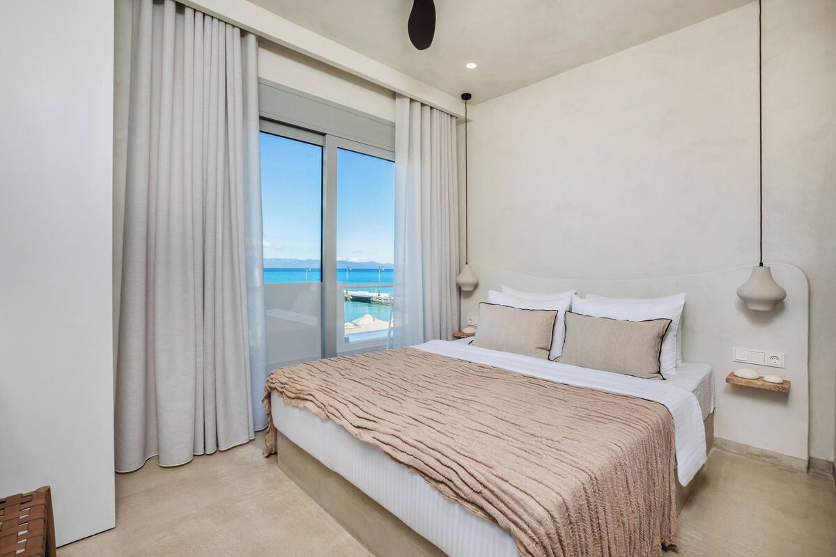 A bedroom with a large bed, a nightstand, a lamp, and a view of the sea.