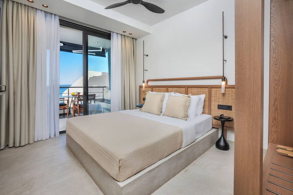 A modern hotel room with a large bed, a balcony with a table and chairs, and a view of the sea.