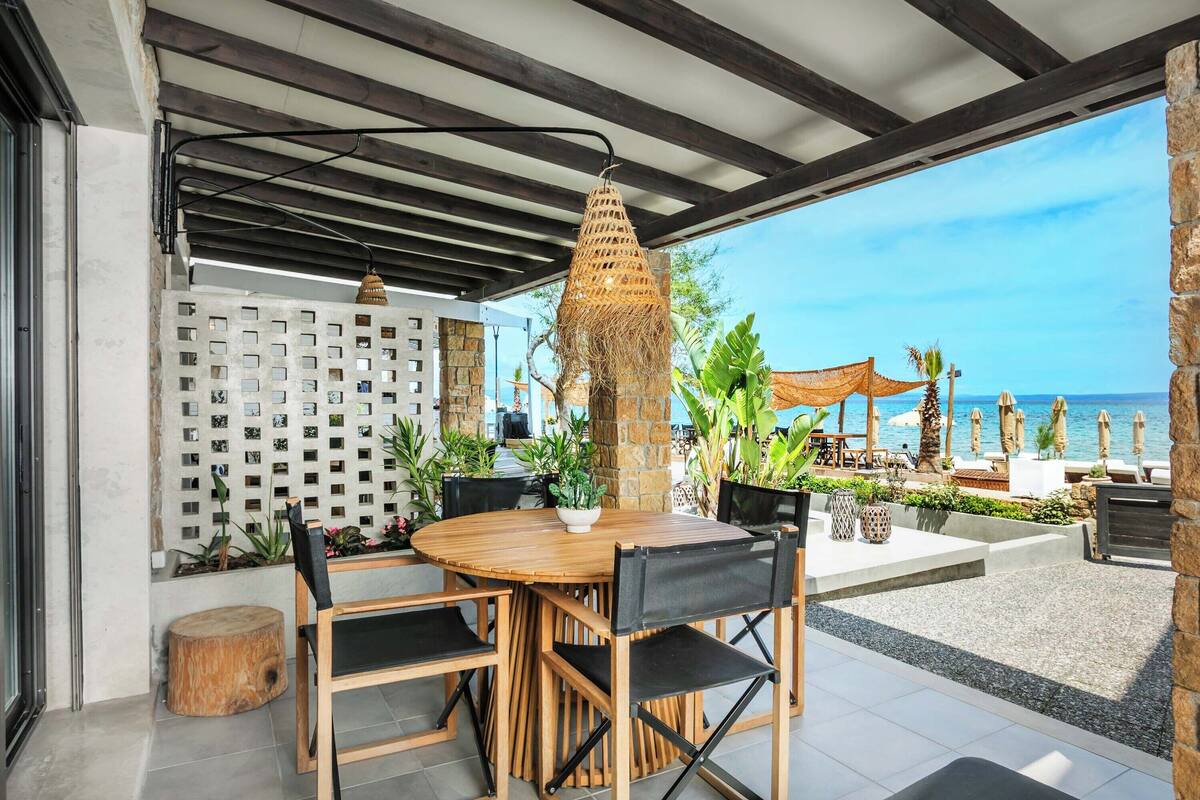 An outdoor dining area with a wooden table and chairs, a hanging light fixture, and a view of the beach.